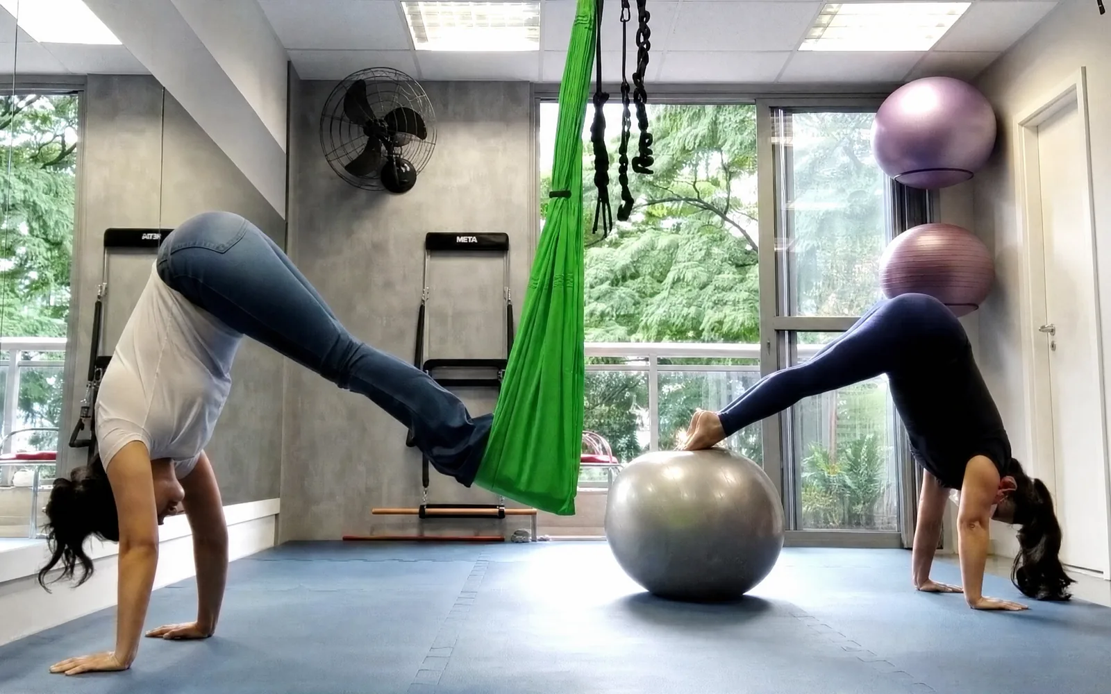Duas alunas em exercício no estúdio da Saúde Urbana, uma apoiada no tecido suspenso e outra na bola, com janelas para as árvores