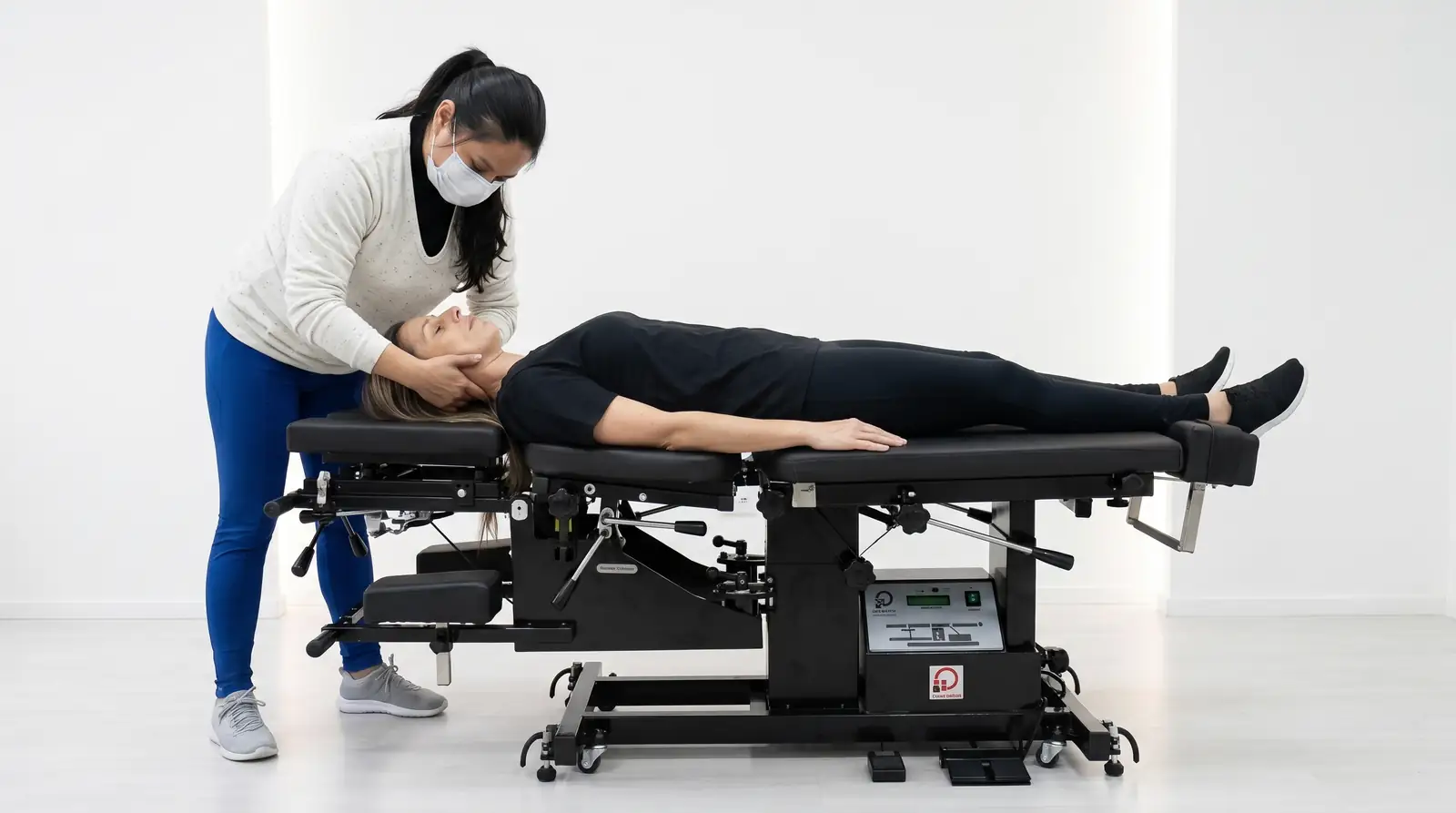 Quiropraxista da Saúde Urbana apoiando com as duas mãos o pescoço de uma paciente deitada, em preparação para o ajuste cervical